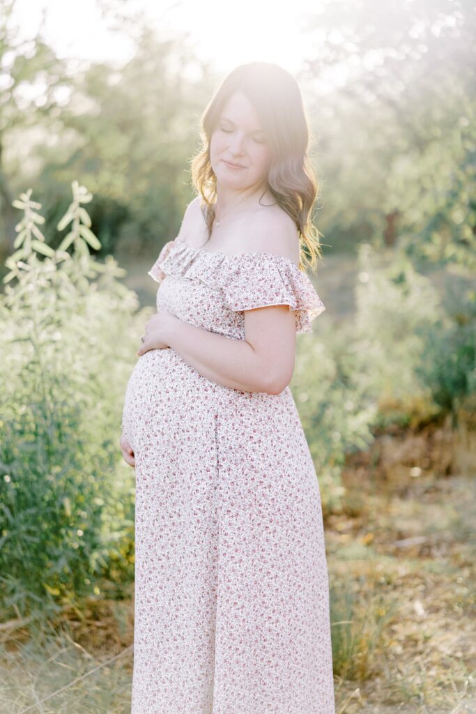 Portrait of a expecting mom standing and holding her baby bump with the sun pouring it behind her