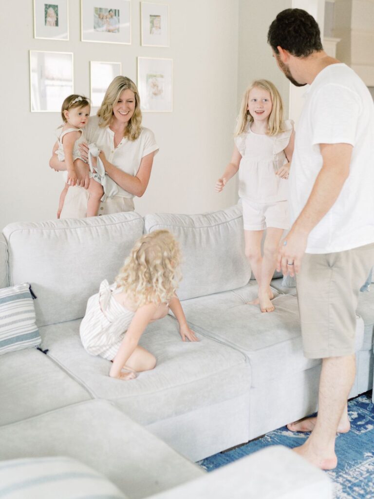 Courtney Houk Photography captures two older daughter jumping on the cough while mom, dad, and baby sister watch