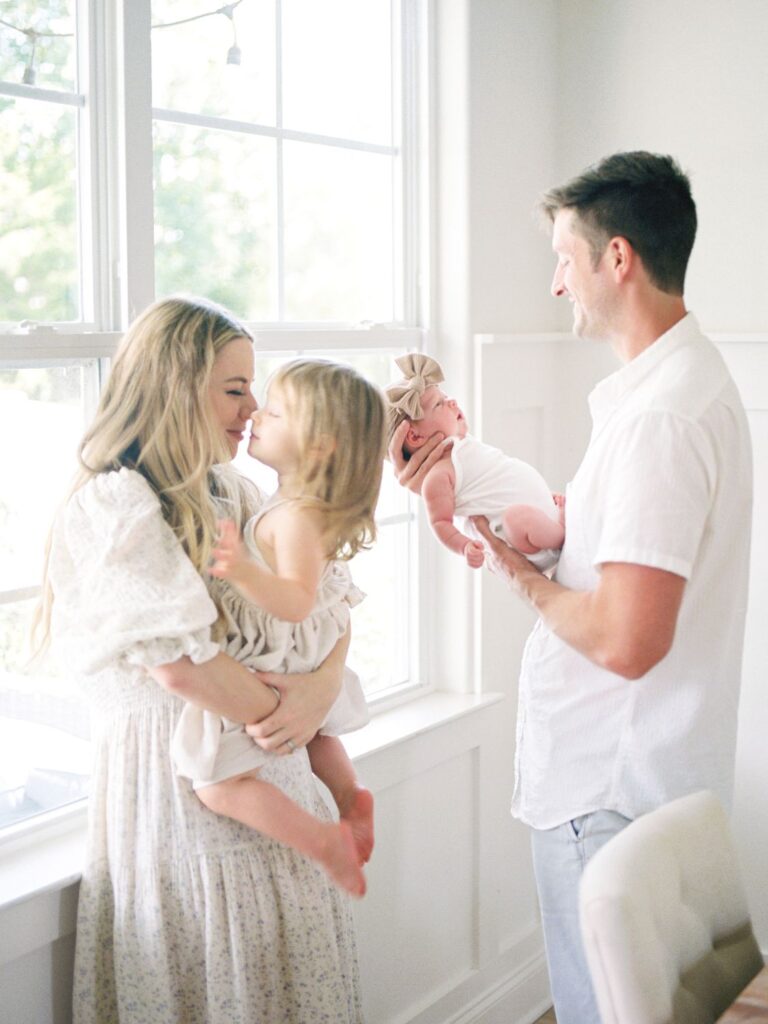 In these in-home Nashville newborn photos, mom does nose to nose with 2 year old daughter while dad holds newborn baby girl in front of him