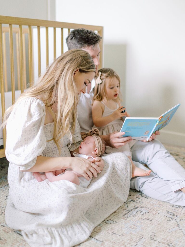 Mom cuddles sleeping newborn baby girl while dad reads book to toddler older sister