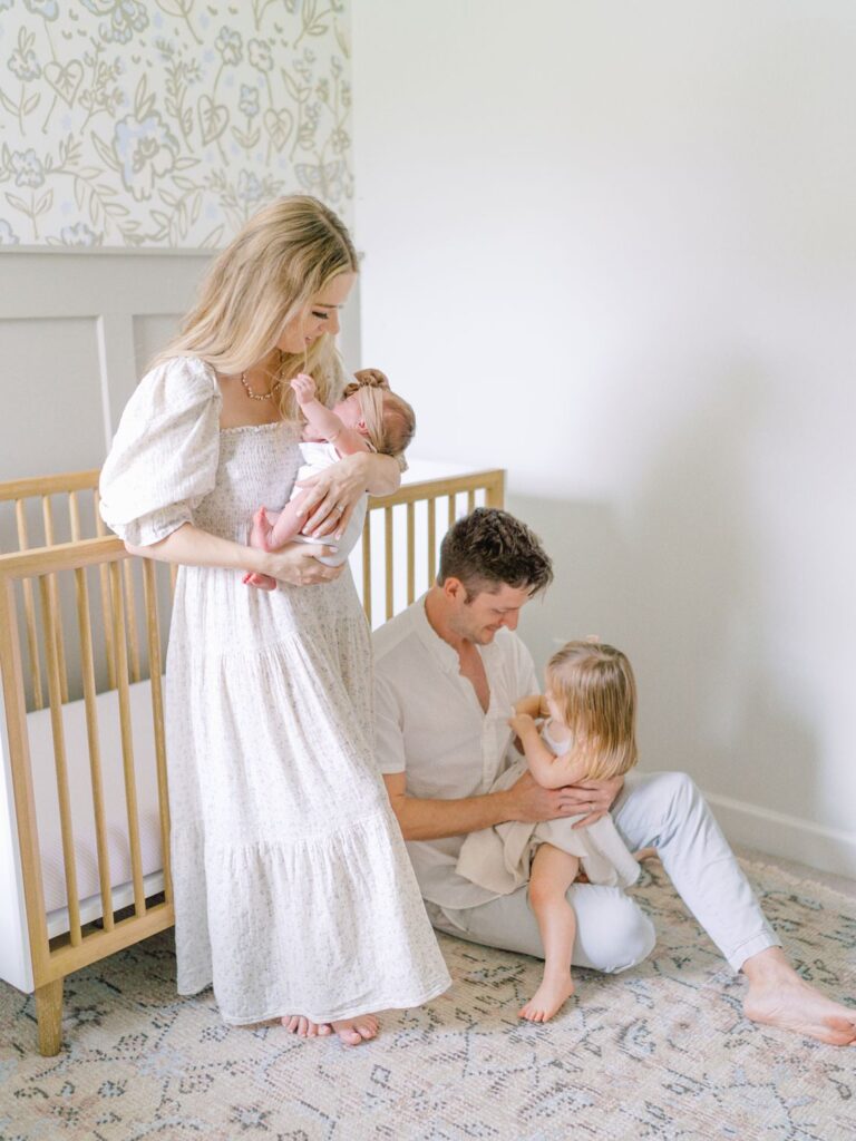 This nursery shot shows mom standing and holding newborn baby girl while dad sits on ground with toddler girl telling her a story