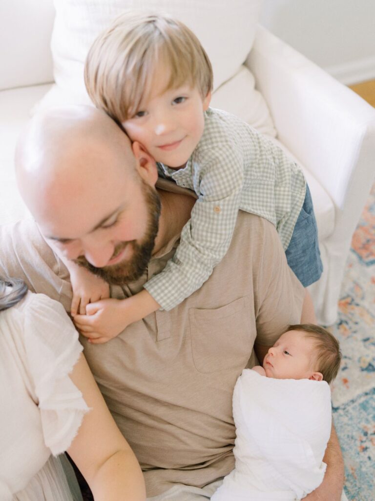 Boy wraps his arms around dad's neck while dad cuddles newborn baby brother in these Nashville newborn photos with siblings