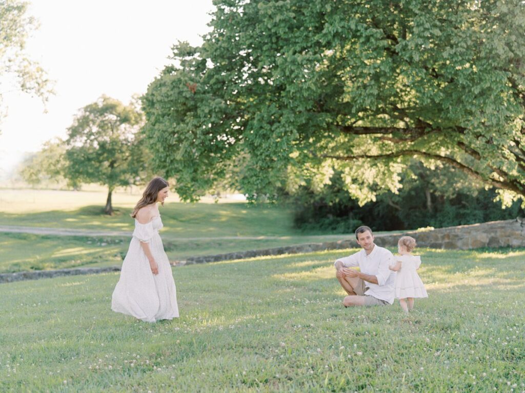 Courtney Houk captured this image of mom walking towards dad kneeling in grassy area and one year old girl next to him at an outdoor Nashville family photos session
