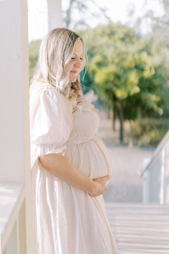 A pregnant mama stands on an all white porch leaning on the railing while holding her belly