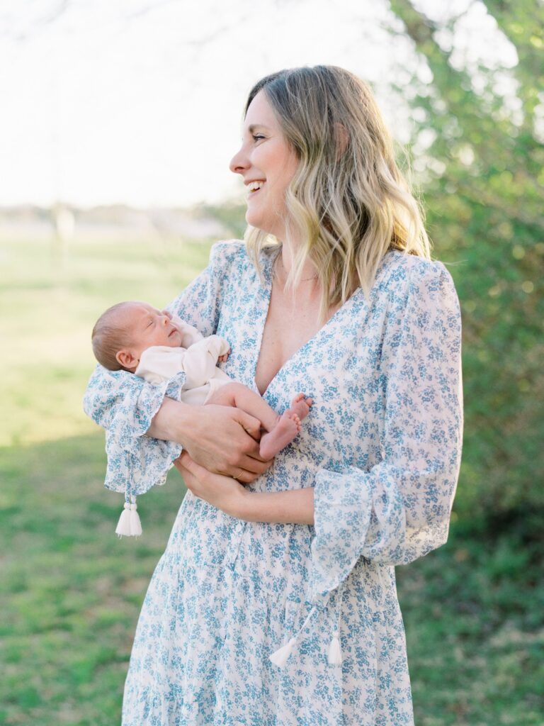 A mama in a long sleeve blue floral dress holding her newborn baby boy while smiling over her left shoulder