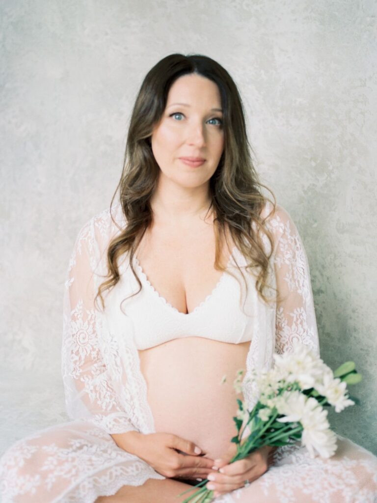 Pregnant mama with a lace shall on sitting on ground holding her bump with flowers in her hand