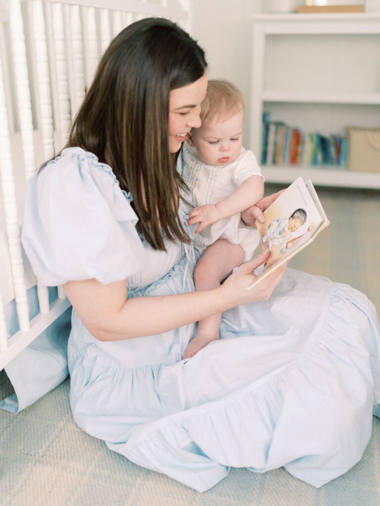 A mama in a blue dress reading a book to her 6 month old baby boy while he is sitting in her lapping snuggling