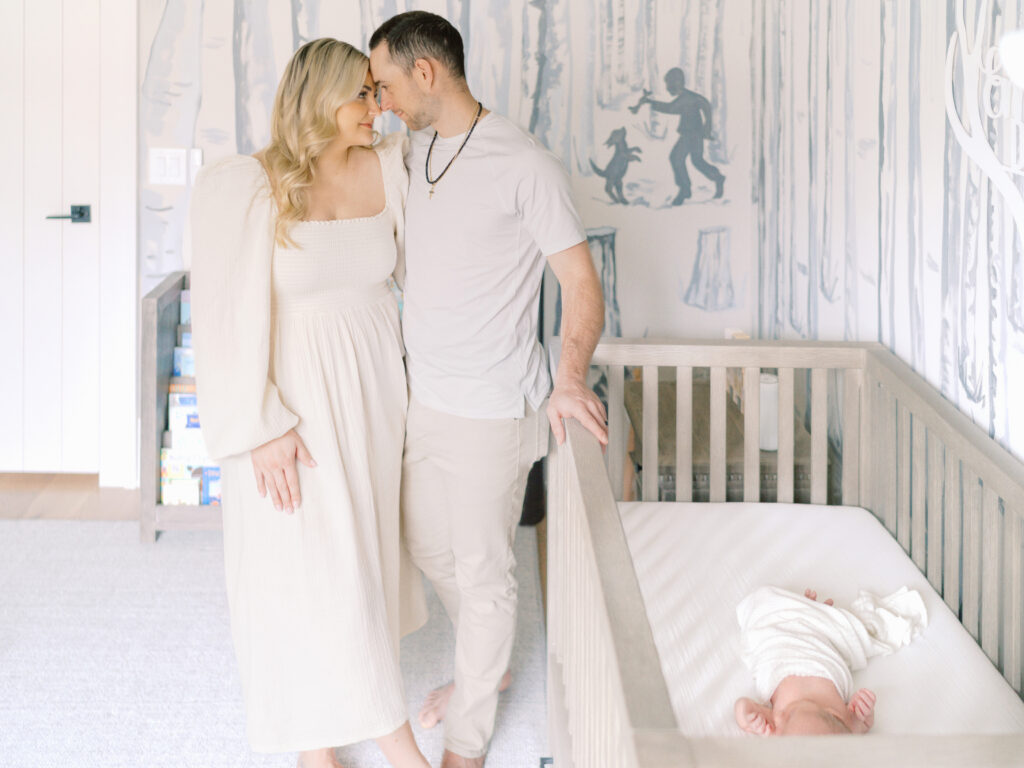 A mom and dad stand at the end of a crib smiling at each other while their baby boy is sleeping