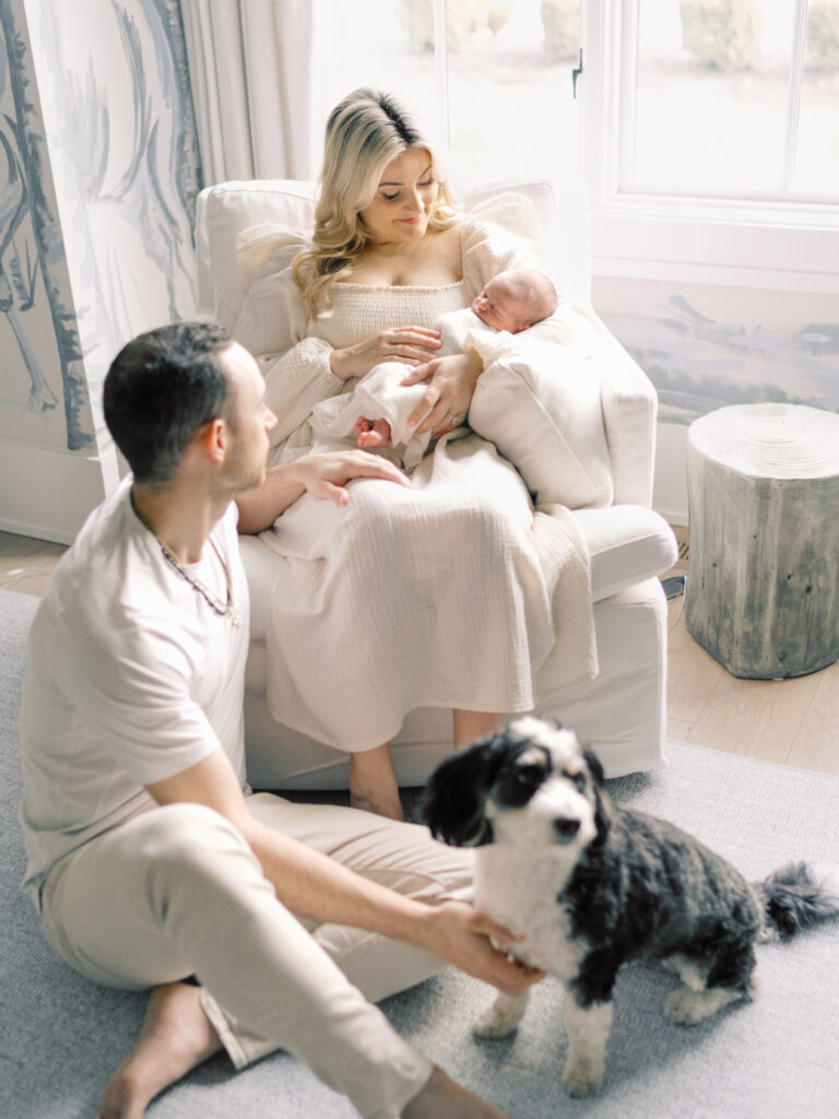 A father sitting on the ground next to a rocker that his wife is rocking in while holding their newborn baby boy