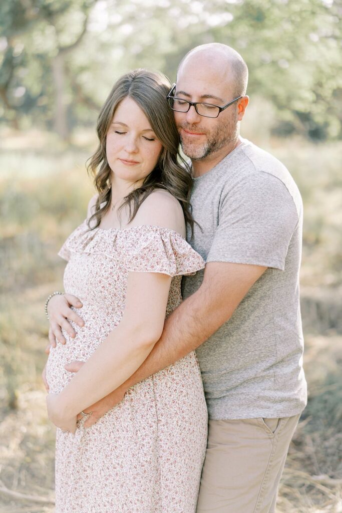 Pregnant mom and dad stand together with hands on mama's baby bump in outdoor maternity session