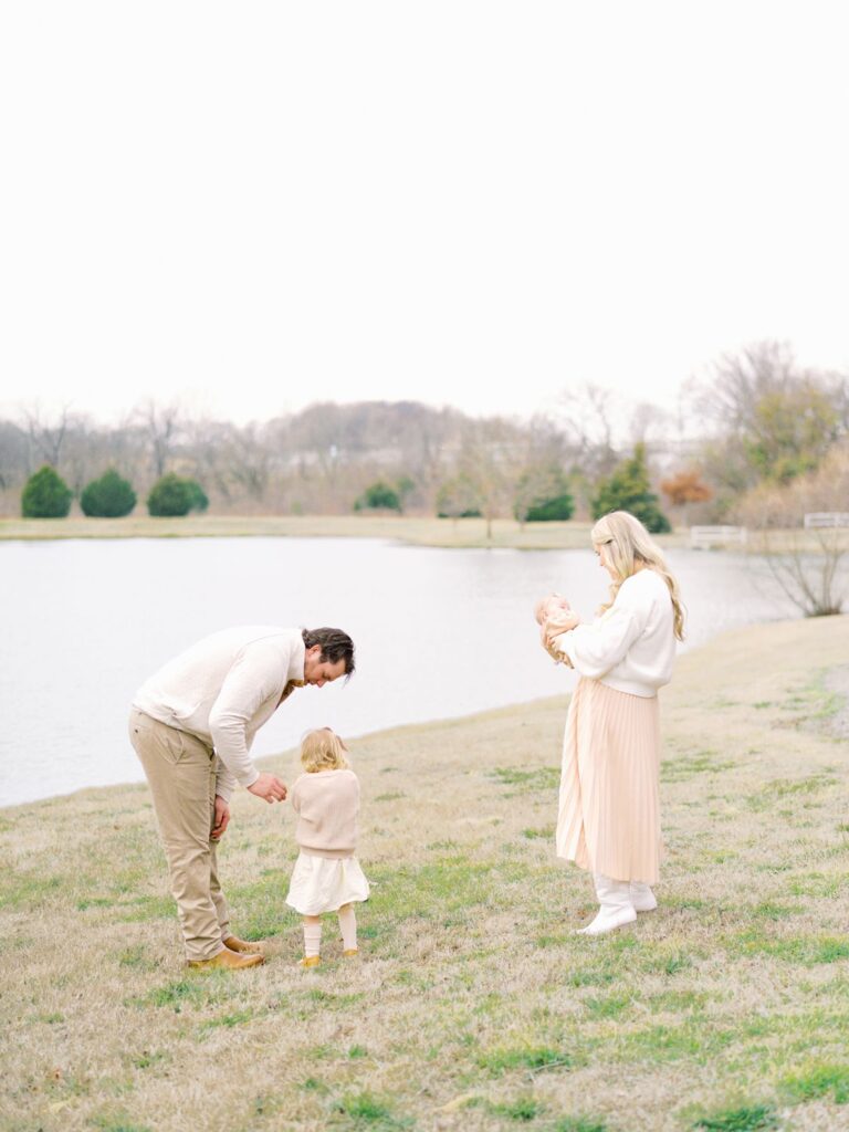 a dad and mama standing in grass next to a pond with their two daughters