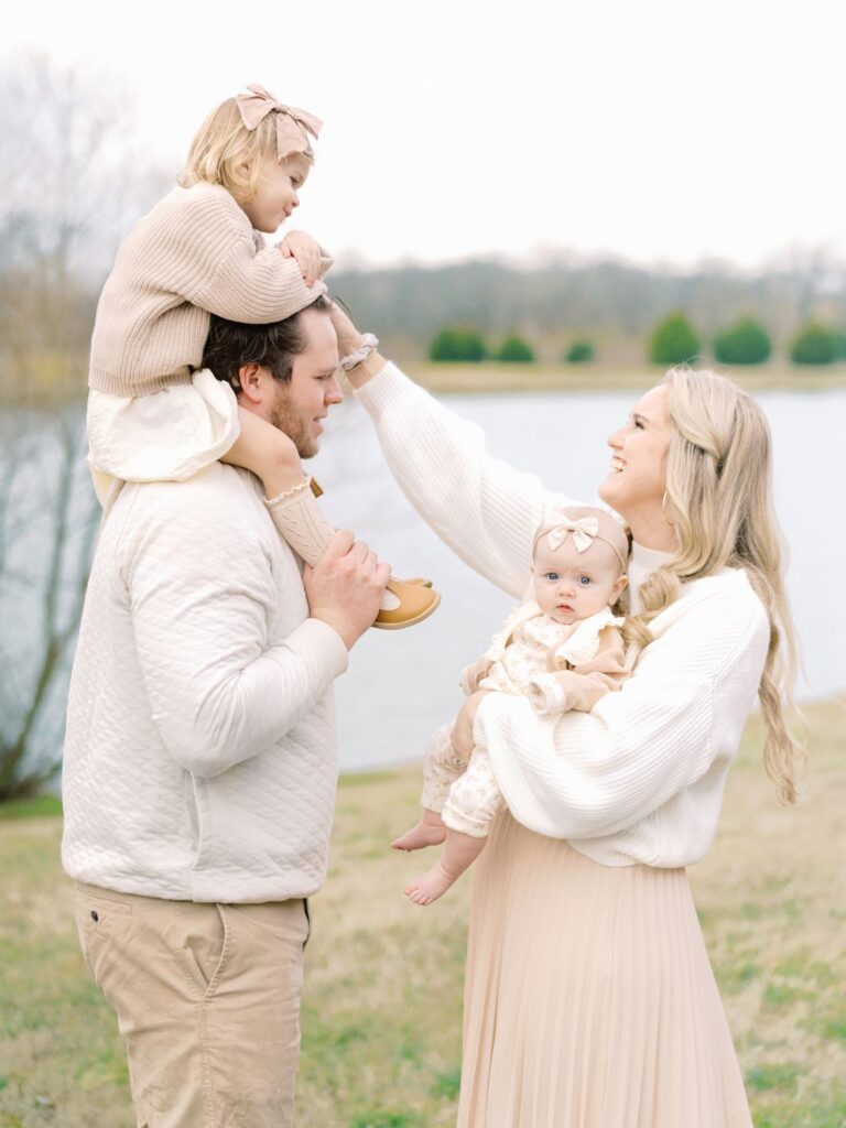 a dad stands with his two year old daughter on his shoulders while mama holds their baby girl 