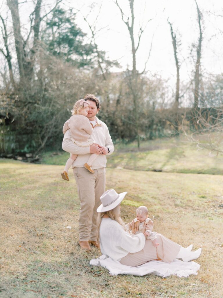 a dad is standing holding his two year old daughter while a mama sits on a blanket with her baby on her lap