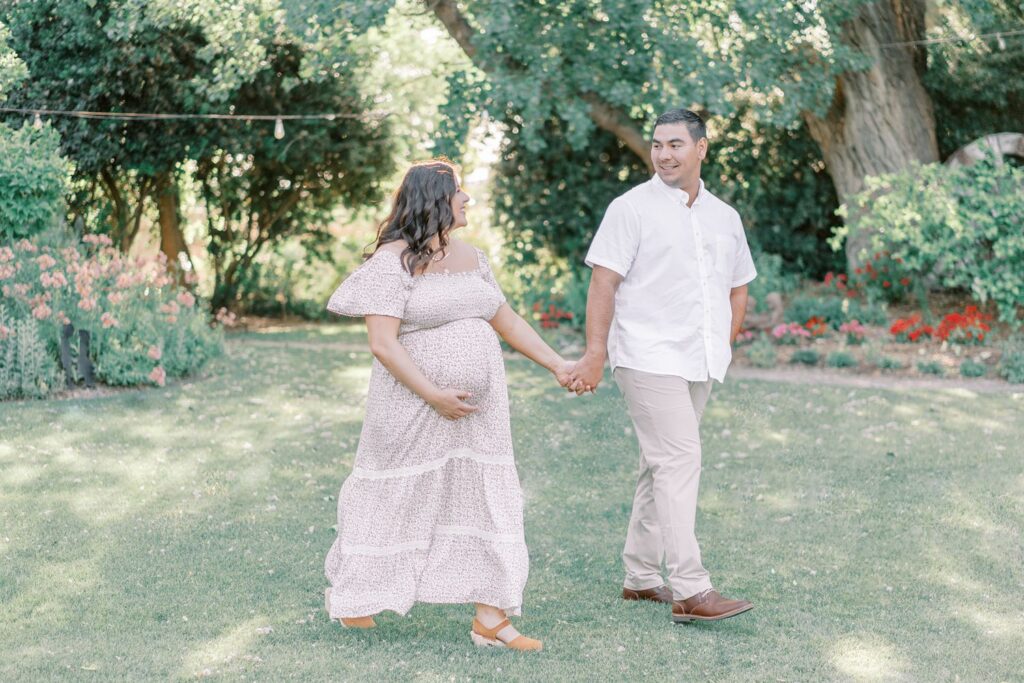 A pregnant mama walks hand in hand with her husband in the grass as they smile at each other