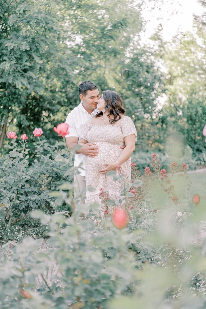 a soon to be dad stands behind his pregnant wife and wraps his arms around her belly in a beautiful flower garden